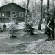 Stadtarchiv Neustadt a. Rbge., ARH Slg. Bartling 4657, Wochenendhaus (Blockhaus) in Kiefernwaldlichtung, Außenansicht Giebelseite, rechts ein Mann bei Verrichtung gärtnerischer Arbeiten, Metel