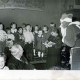 Stadtarchiv Neustadt a. Rbge., ARH Slg. Bartling 619, Weihnachtsfeier für Vorschulkinder, Gruppe von Kindern auf einer Bühne, davor rechts der Weihnachtsmann, über den Kindern an der Wand ein Brett mit den Daten der Baugeschichte des Vereinsheims, Stöckendrebber